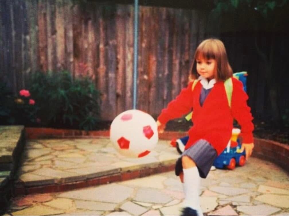 A young Rebecca in school uniform kicking a football in a backyard