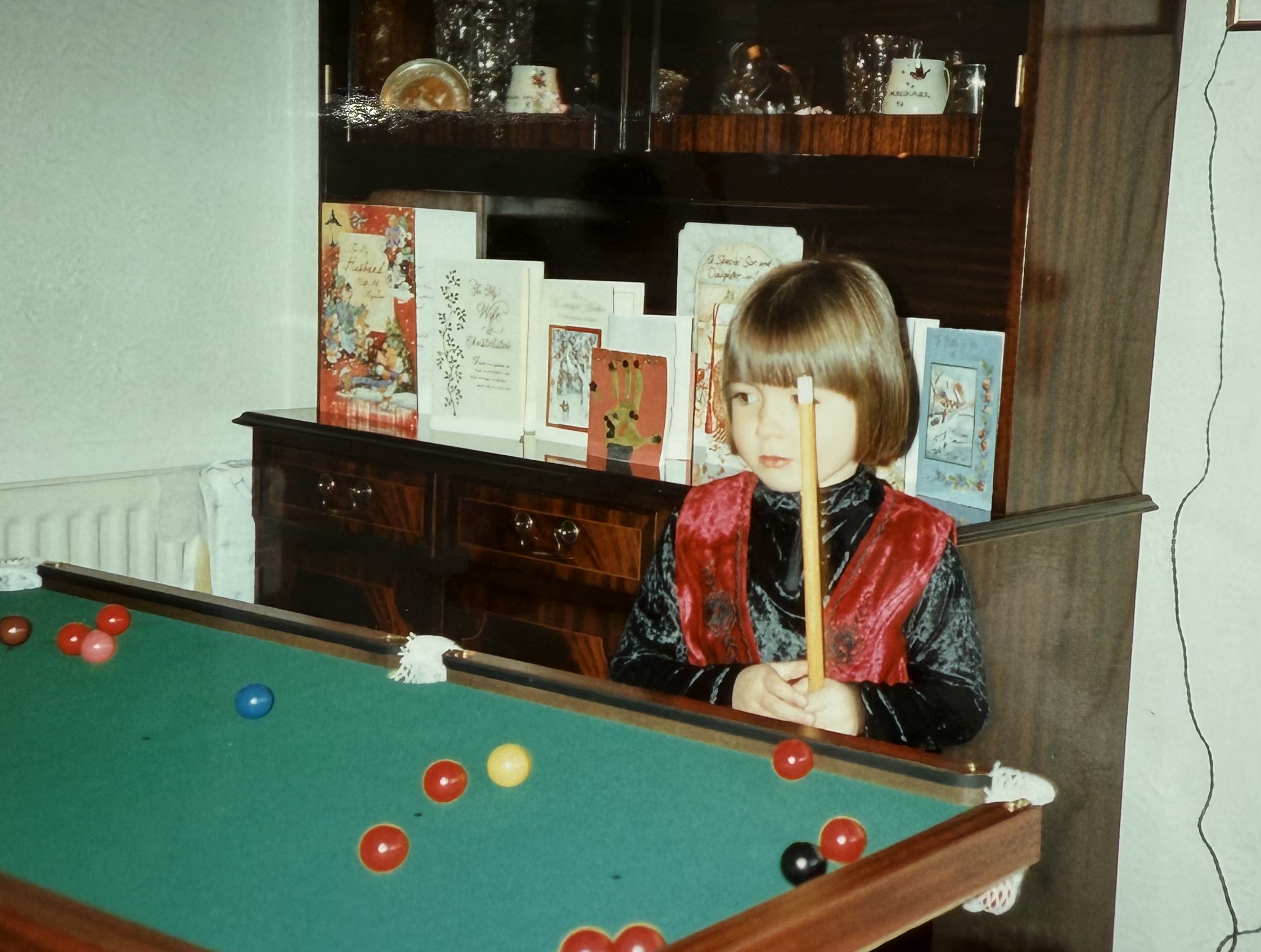 A young Rebecca holding a pool cue beside a tabletop snooker table