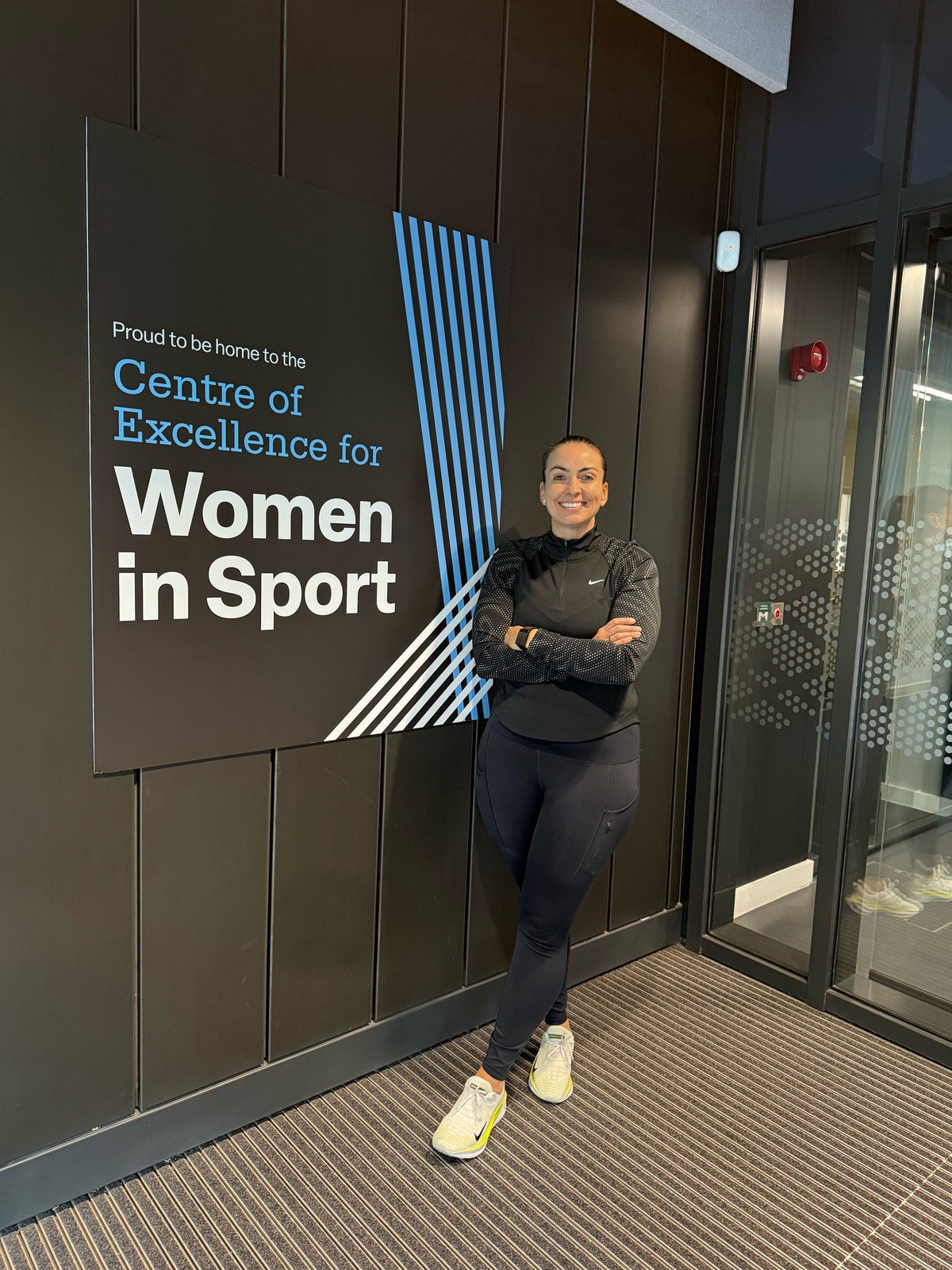 Rebecca standing with arms crossed beside a sign reading 'Proud to be home to the Centre of Excellence for Women in Sport'