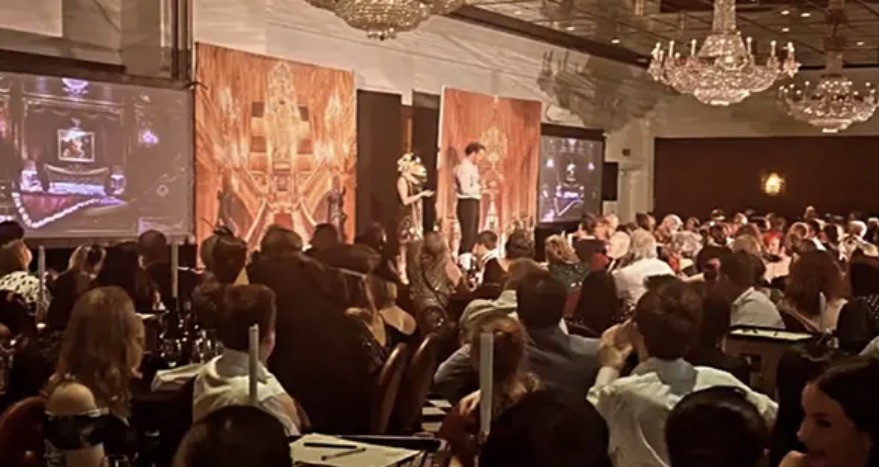 A gala dinner event in a grand ballroom, with guests seated at tables facing a stage under crystal chandeliers