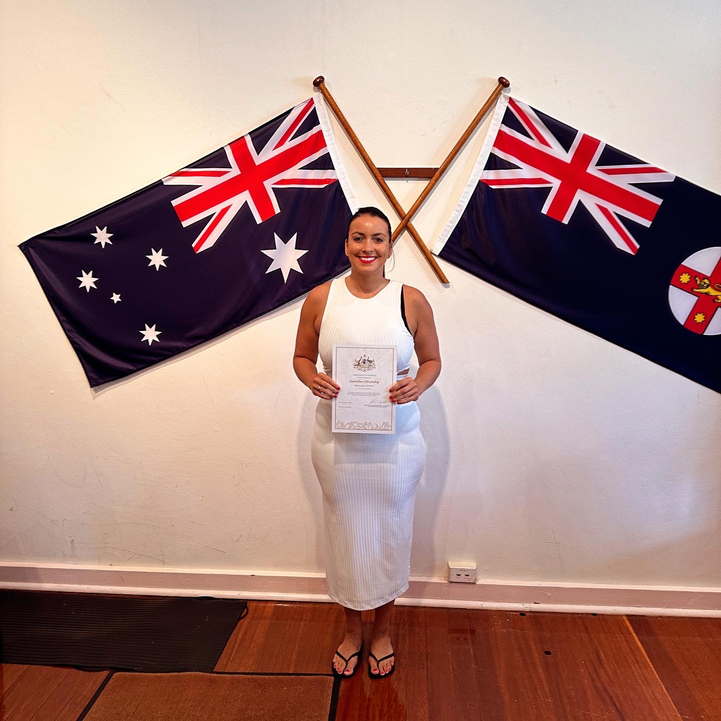 Rebecca smiling and holding her Australian Citizenship certificate in front of the Australian and New South Wales flags